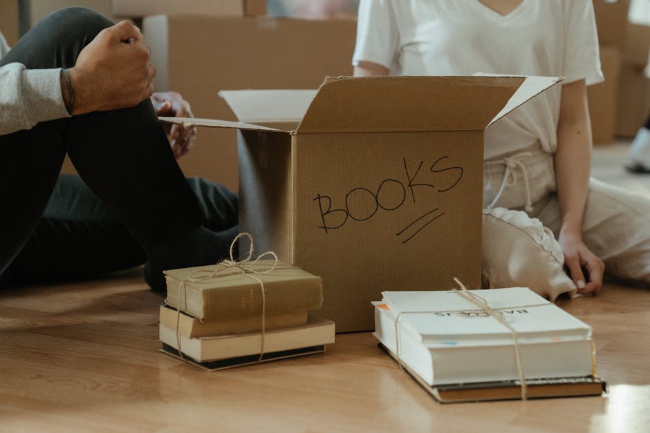 A woman and a man are inside a residential property with a plain light-colored wall in the background, engaged in packing belongings for a house move. The woman, wearing a white top, is holding and pa