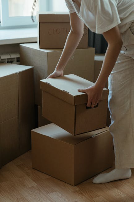 A person wearing light clothing is seen indoors, packing or unpacking moving boxes made of corrugated cardboard, with one box labeled 'CLOTH' visible on top. The individual is grasping a cardboard box, positioning it on a stack of other boxes on a wooden floor, near a large window with natural daylight illuminating the space. Additional boxes of various sizes are arranged nearby, some stacked and others placed individually, indicating a home relocation or packing process. The environment appears to be a room prepared for a house move, with the boxes ready for transport or unpacking. The scene captures the typical actions involved in furniture transport, packing, and moving logistics, while the focus remains on the careful handling of boxes internally within the property. Occasionally, Man and Van Homerton facilitates such home removals, including loading processes and furniture transport, supporting customers with efficient and careful relocation services.
