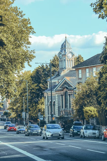 A street scene in Homerton with several parked cars along the curb and a professional man with a moving trolley loaded with cardboard boxes and wrapped furniture inside a property’s doorway. The background features a historic clock tower with a spire, surrounded by trees, residential buildings, and a clear blue sky. The setting indicates a home relocation or packing and moving process, with the focus on the loading area outside the property where furniture and boxes are prepared for transport. Natural daylight illuminates the scene, highlighting the details of the furniture, packaging materials, and the urban environment, aligning with house removals services offered by Man and Van Homerton.