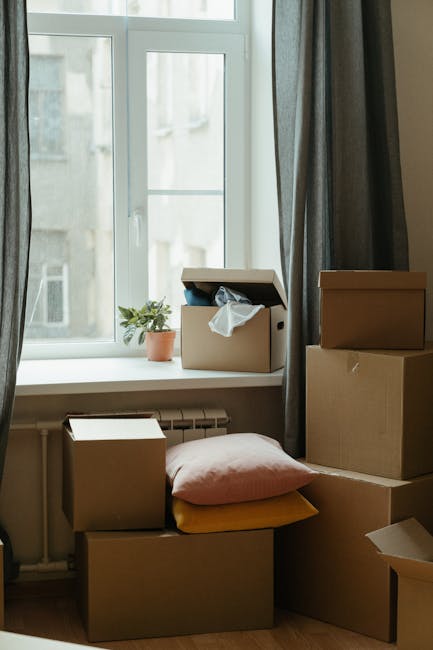 A person wearing light clothing is seen indoors, packing or unpacking moving boxes made of corrugated cardboard, with one box labeled 'CLOTH' visible on top. The individual is grasping a cardboard box, positioning it on a stack of other boxes on a wooden floor, near a large window with natural daylight illuminating the space. Additional boxes of various sizes are arranged nearby, some stacked and others placed individually, indicating a home relocation or packing process. The environment appears to be a room prepared for a house move, with the boxes ready for transport or unpacking. The scene captures the typical actions involved in furniture transport, packing, and moving logistics, while the focus remains on the careful handling of boxes internally within the property. Occasionally, Man and Van Homerton facilitates such home removals, including loading processes and furniture transport, supporting customers with efficient and careful relocation services.