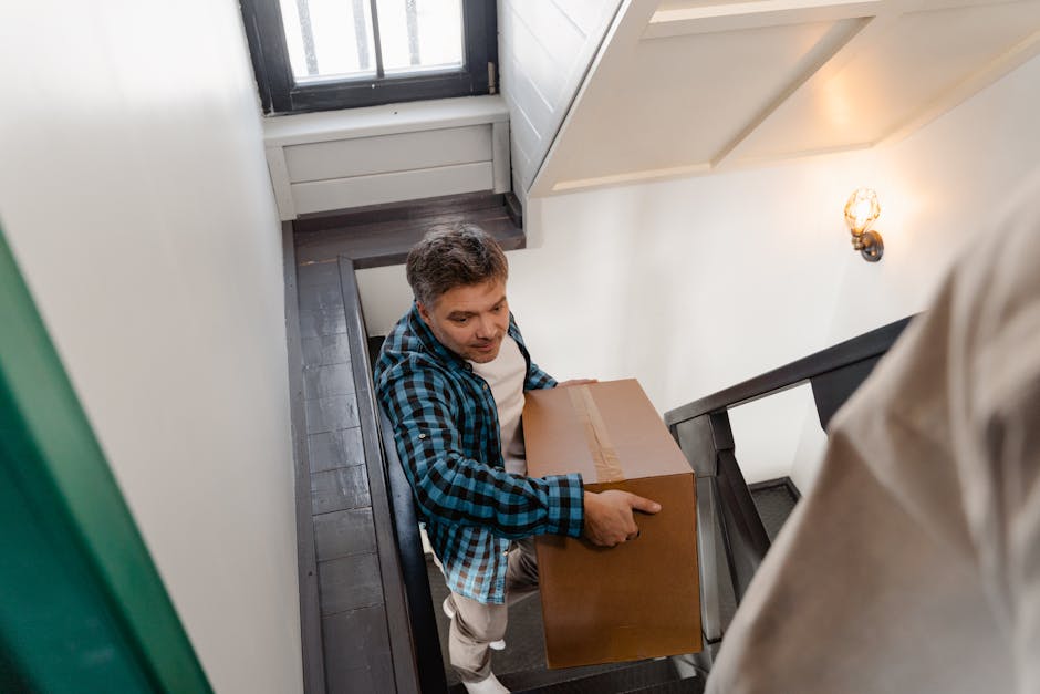 A man wearing a blue and black checkered shirt is carrying a cardboard box during a home relocation, navigating a narrow staircase inside a property. The stairway has dark wooden treads and black railings, with white walls on either side. Natural light from a nearby window illuminates the area. The man appears focused on the moving process, which involves carefully lifting and guiding the box down the staircase as part of furniture transport and packing and moving activities. In the background, there is a small wall-mounted sconce with a warm light bulb, and the adjacent wall features a partially visible door or window frame. This scene is part of a professional removal service by Man and Van Homerton, specializing in house removals and relocation logistics, including narrow access moves on Homerton High Street.