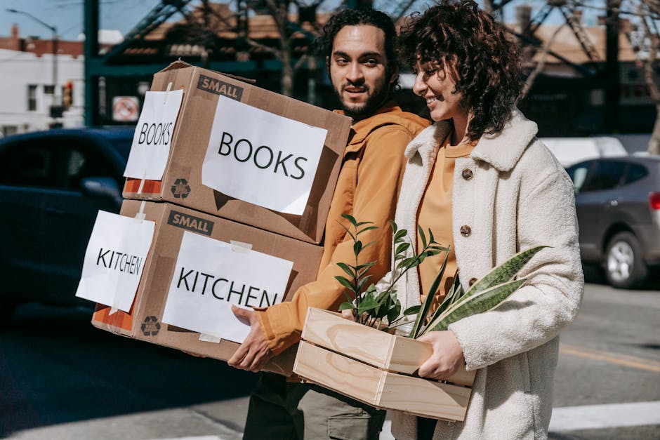 Two people, a man and a woman, are outdoors on a street with parked cars and buildings in the background, involved in a home relocation process. The man, with dark hair and a beard, is holding two cardboard boxes labeled 'BOOKS' and 'KITCHEN' in black text, which are secured with packing tape and feature labels indicating contents for furniture transport. The woman, with curly hair, is smiling and holding a wooden tray with several potted plants, including leafy green foliage. Both are dressed in casual clothing, suitable for moving day. The scene captures the loading or unloading phase of a house removal, with natural daylight illuminating the setting. This image is relevant to packing and moving services, showcasing the process of organizing belongings for a move, and reflects the logistics involved in house removals, particularly when navigating narrow or constrained access areas as described in the Homerton High Street removals guide for narrow access moves. Man and Van Homerton official services are implied through the context.