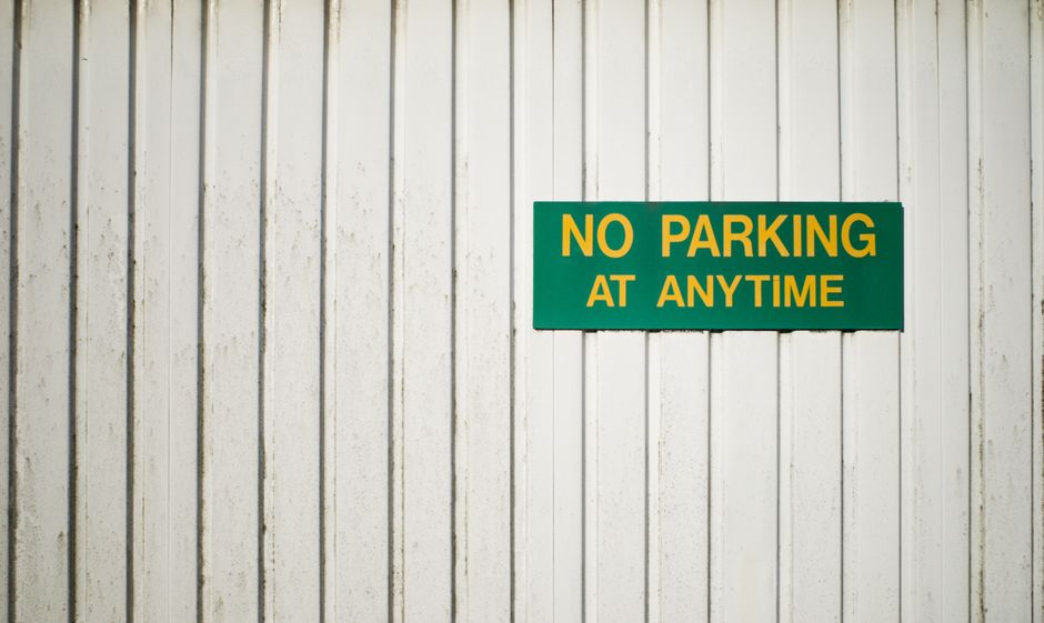 A close-up view of a white wooden wall with vertical paneling, featuring a rectangular green sign with yellow text that reads 'NO PARKING AT ANYTIME'. The sign is mounted on the wall, indicating parki
