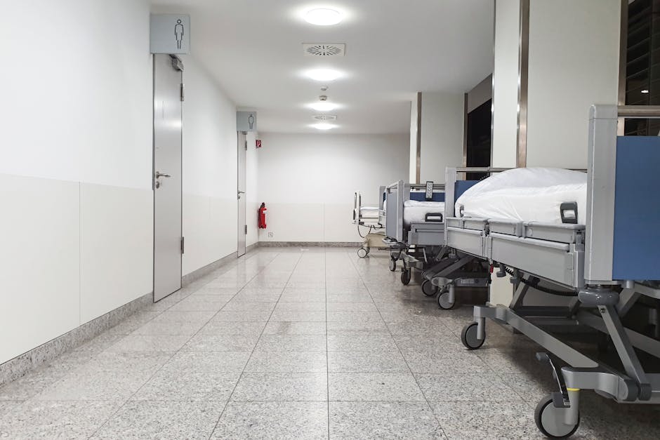 An interior view of a hospital corridor at Homerton University Hospital, with plain white walls and a light wooden handrail running along the left side. The floor is covered with shiny, light-colored