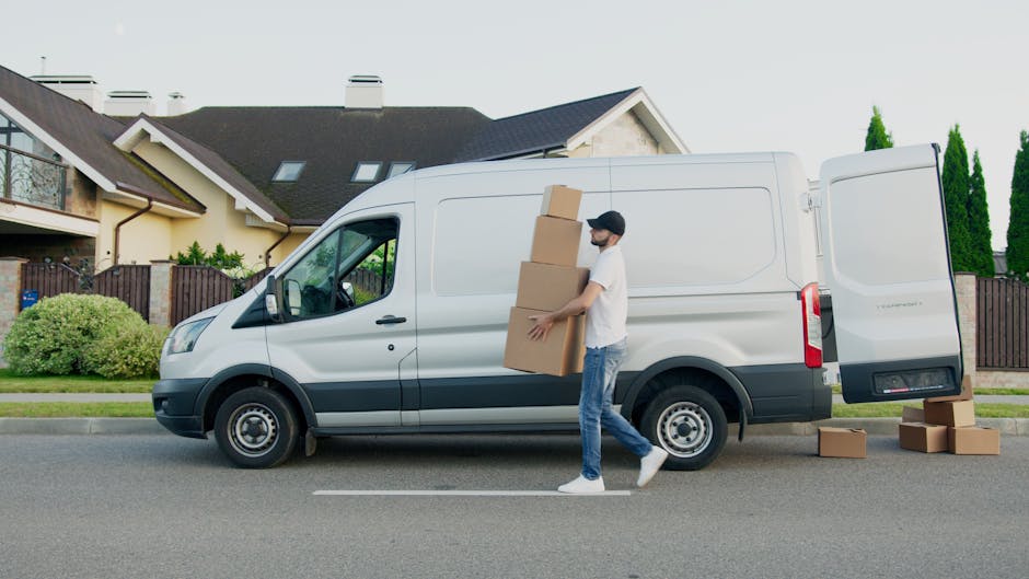 Two movers from Man and Van Homerton are engaged in a home relocation process, transferring a large cardboard box from the driveway into a white commercial van. The scene takes place outdoors during d
