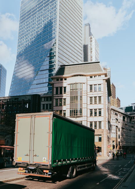 A large green transport truck with a closed cargo area is parked on a city street in front of a mix of modern and historic buildings, including a tall glass skyscraper and an older stone-fronted struc